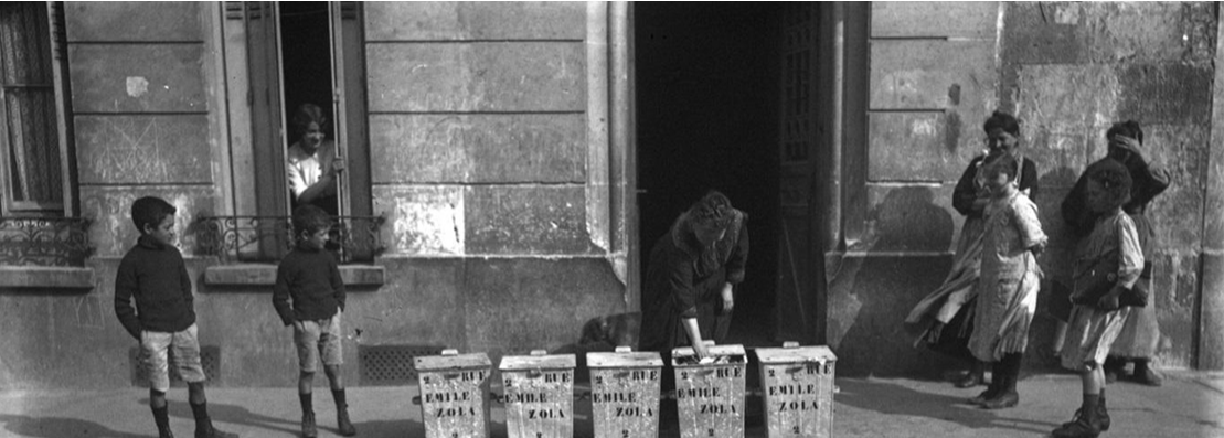 Scène de rue historique montrant des habitants devant des poubelles alignées portant l’inscription « Rue Emile Zola »