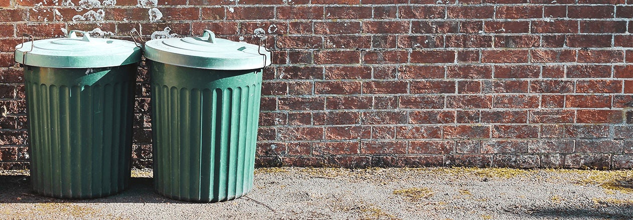 Deux poubelles vertes métalliques sont alignées sur le trottoir devant un mur en briques rouges, illustrant la gestion des déchets urbains