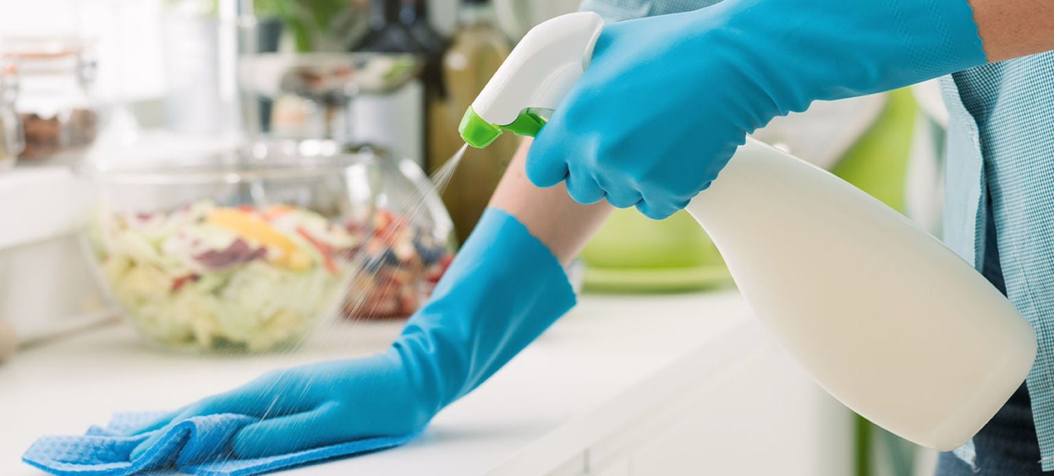 Mains gantées de bleu essuyant un plan de travail de cuisine avec un chiffon et un vaporisateur