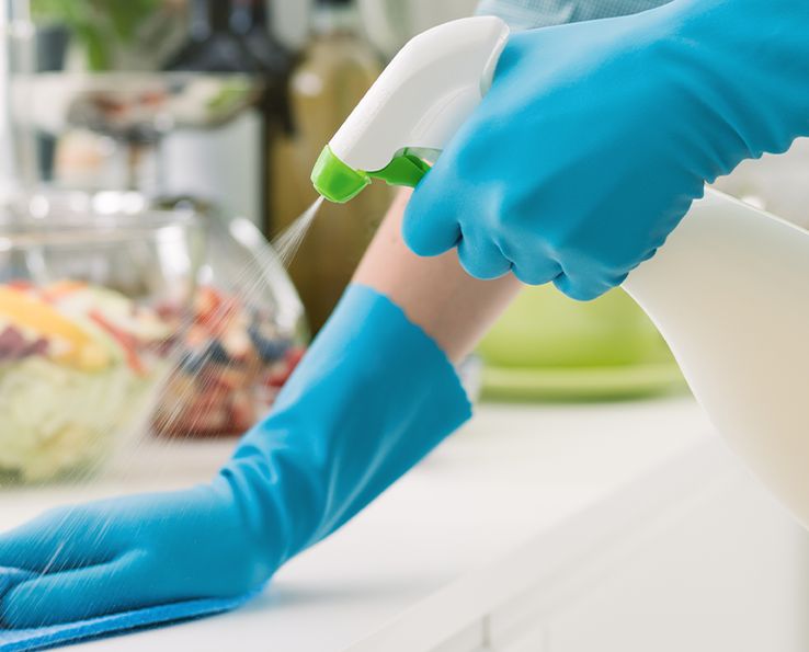 Mains gantées de bleu essuyant un plan de travail de cuisine avec un chiffon et un vaporisateur